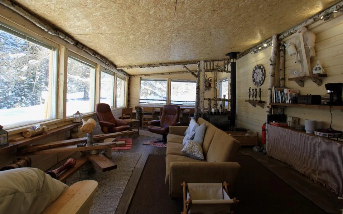 Intérieur du bus « Into The Wild » : longues baies vitrées sur la forêt enneigée, poêle et fauteuils