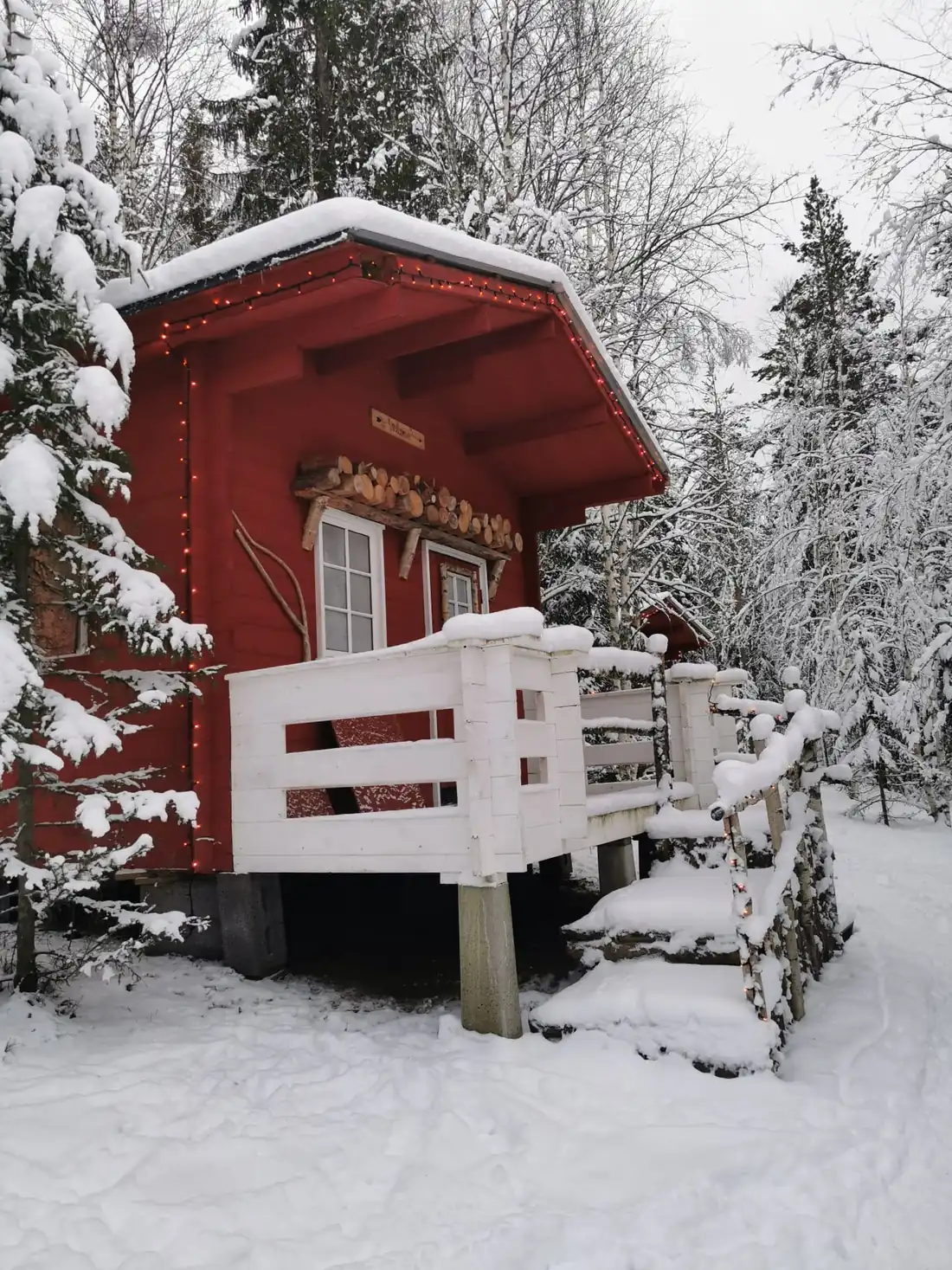 Petit chalet rouge sur pilotis et sa terrasse enneigée, entouré de sapins chargés de neige