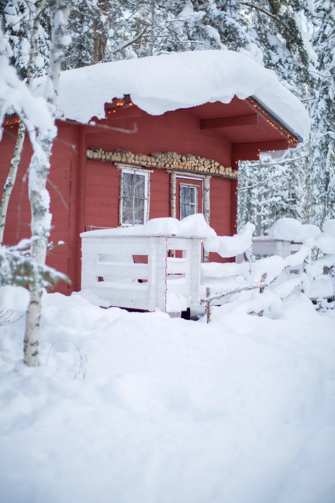 Petit chalet-sauna rouge sous une épaisse neige, guirlandes lumineuses et forêt givrée