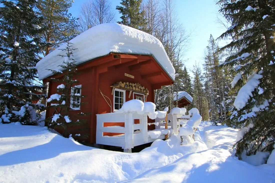 Chalet rouge coiffé d'un épais manteau de neige, sa terrasse et sa balustrade blanche au cœur de la forêt