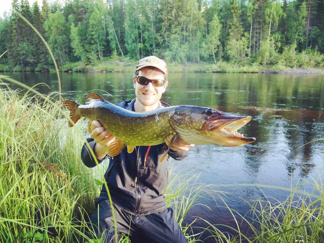 Pêcheur souriant brandissant un gros brochet au bord d'une rivière bordée de forêt verte