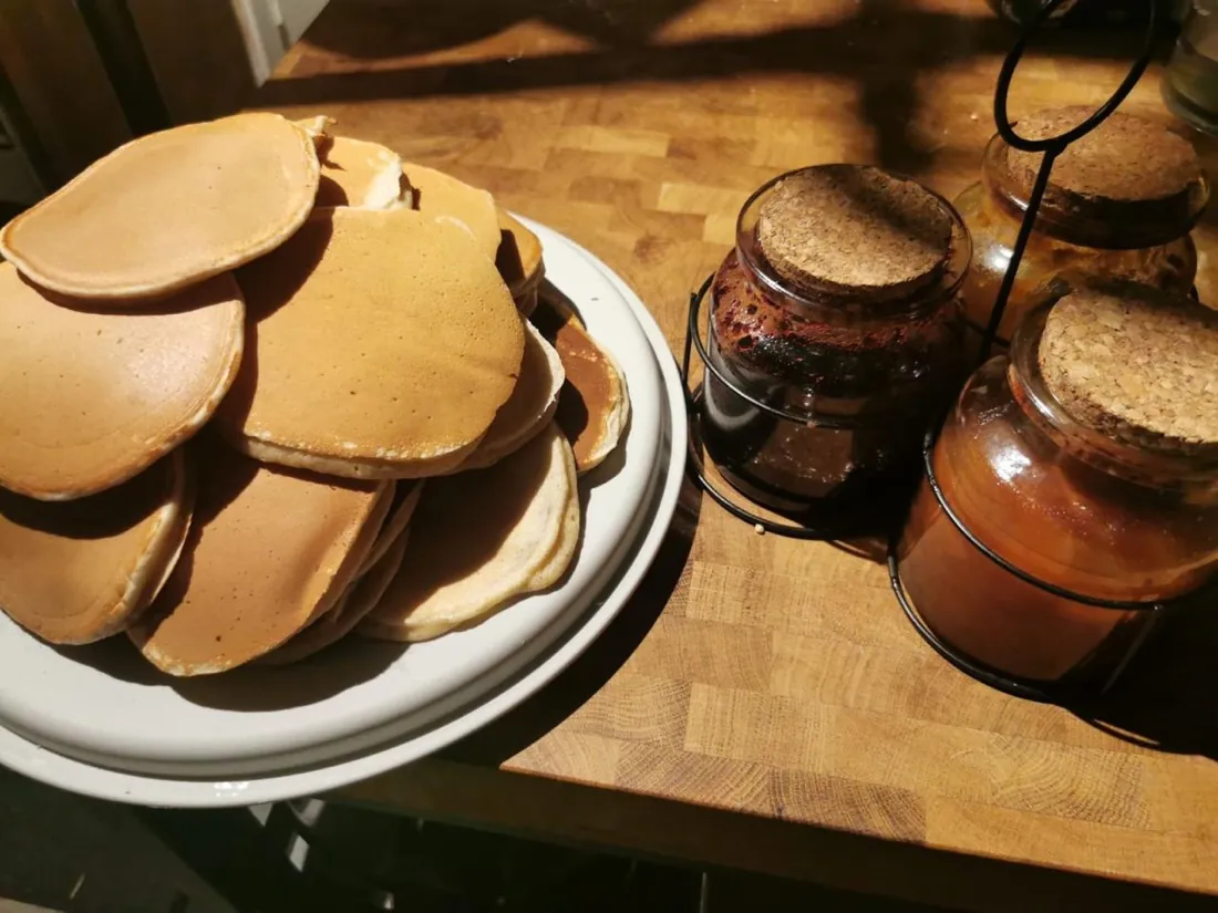 Pile de pancakes maison sur un plat blanc, à côté de pots de confiture et de sirop